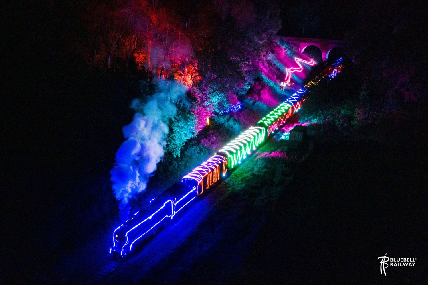 A steam train lit up with multicoloured lights on a railway track in the dark. The colours are bright and the steam is illuminated.