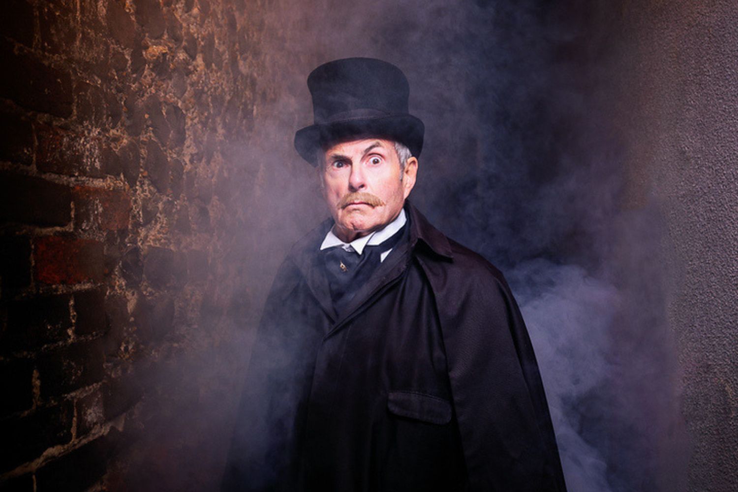 A man is pictured wearing all black with a top hat, raising an eyebrow. He has a grey moustache and grey hair. He is pictured outside, standing in front of bricks, and there is smoke.